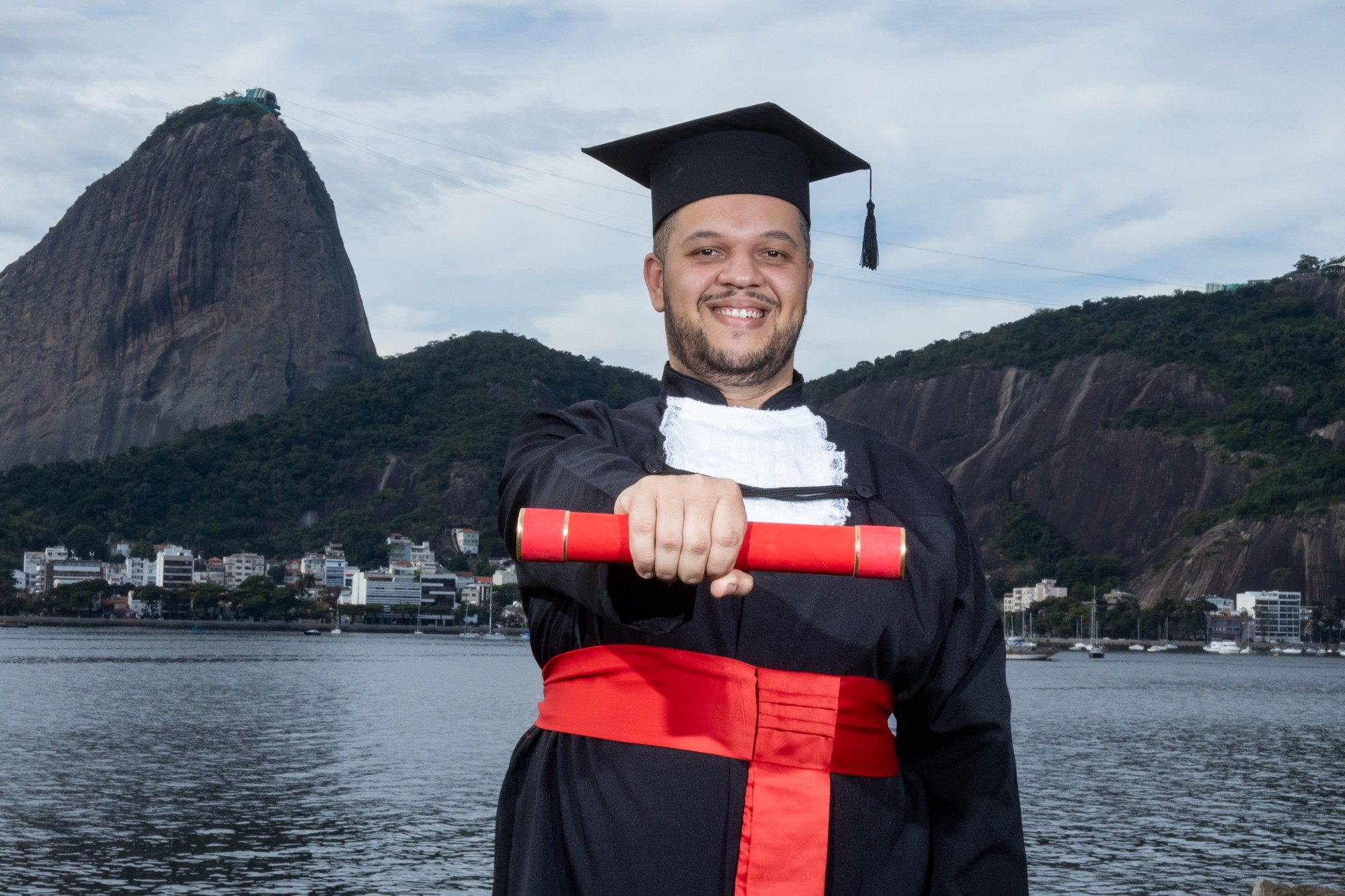 Diego Moutinho de beca e capelo, segurando o canudo do diploma, com o Pão de Açúcar ao fundo